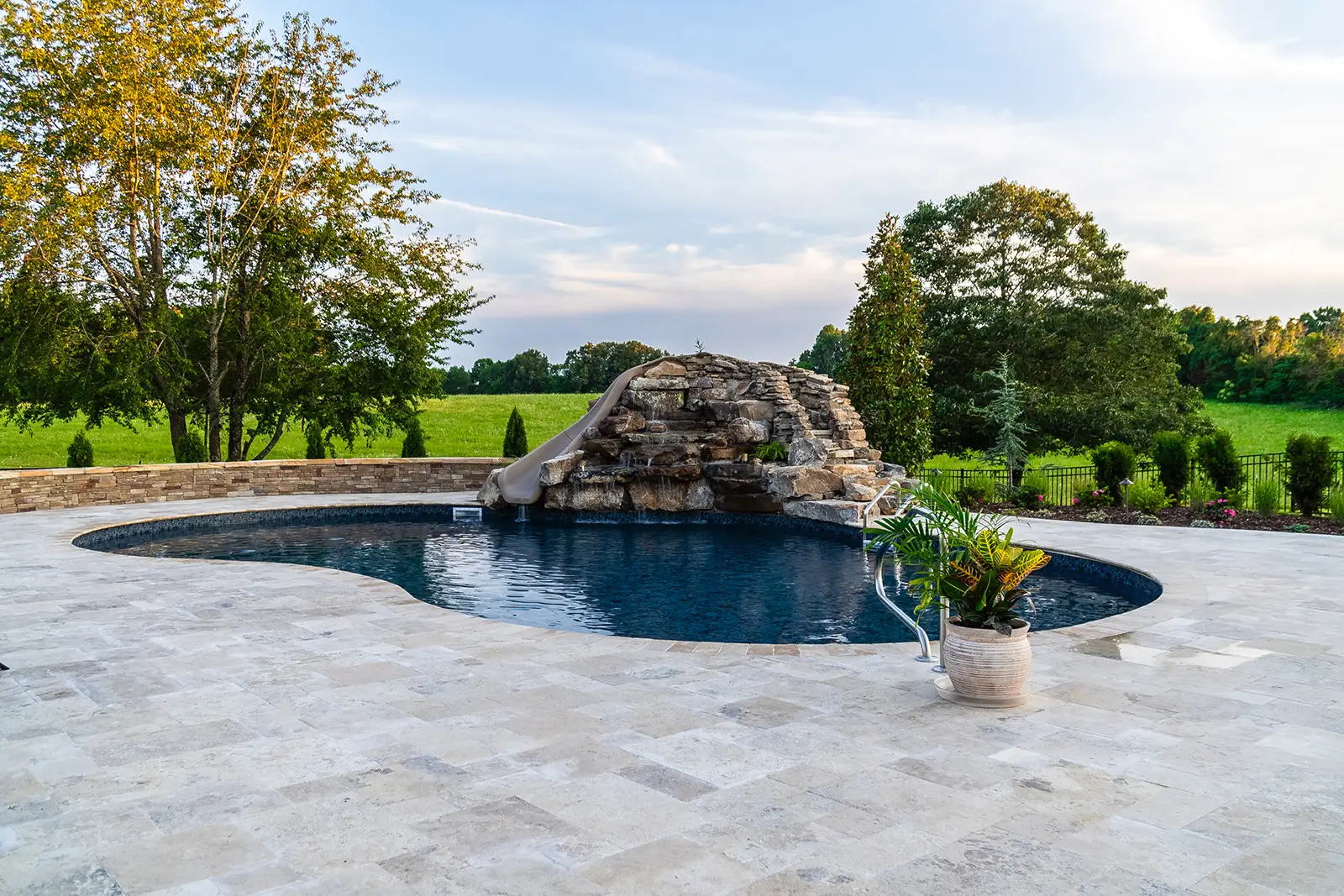 A kidney-shaped pool with a waterfall feature is surrounded by a stone deck. There are trees and a grassy field in the background.